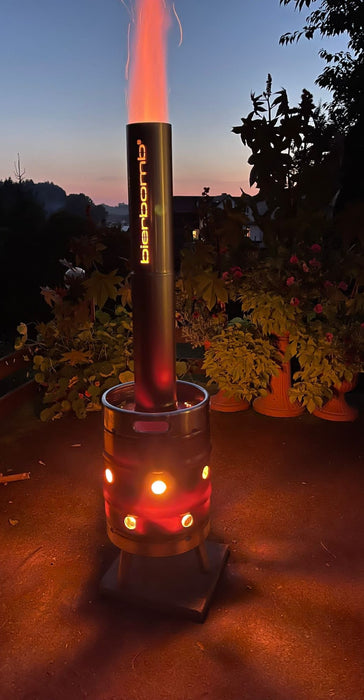 bierbomb® bei Dämmerung auf einer Terrasse, hohe Flamme über dem Rohr, Blumen und Häuser im Hintergrund