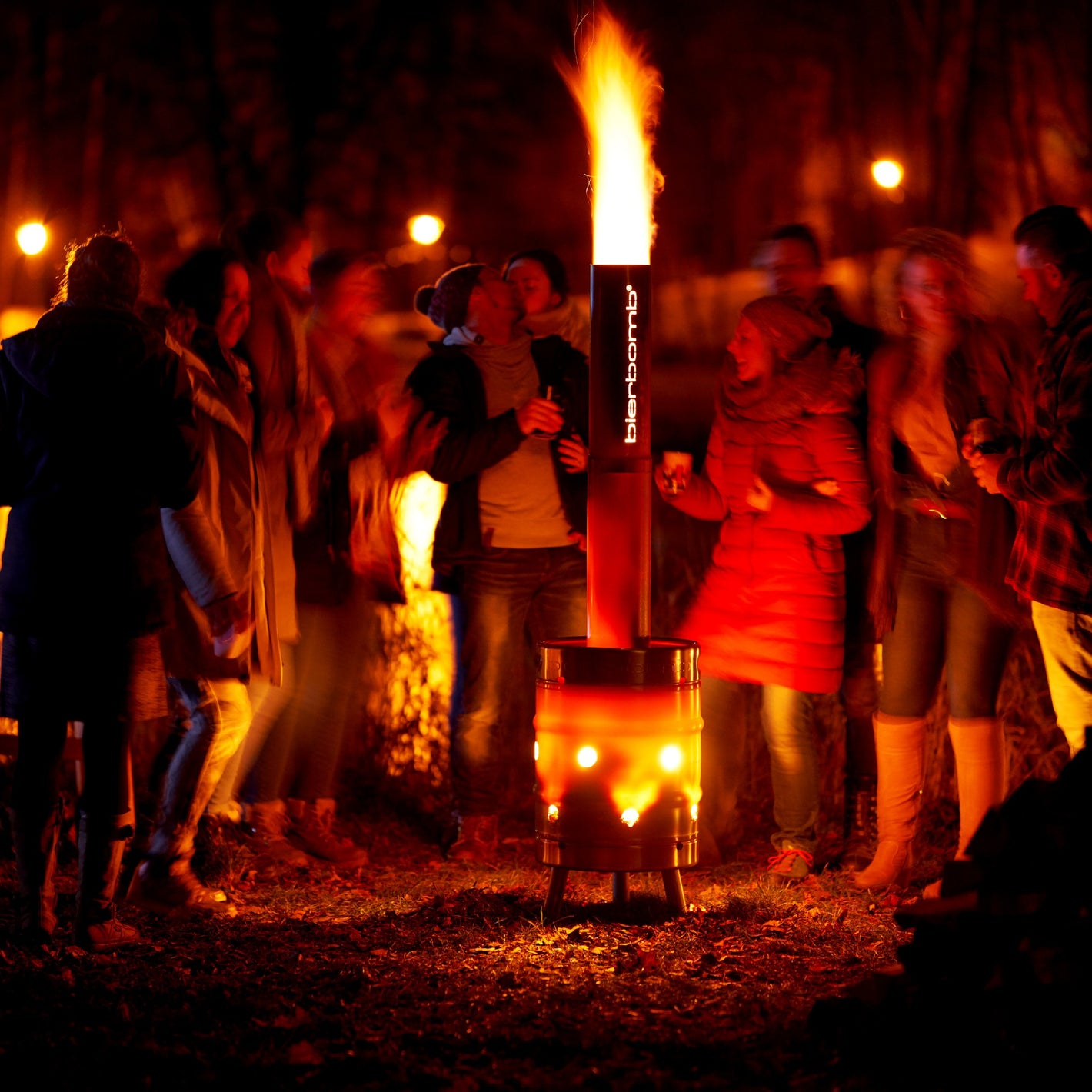 Gruppe von Gaesten steht bei Nacht um die brennende bierbomb Feuertonne, Flamme schlaegt aus dem Schornstein und die Luftloecher gluehen orange