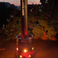 bierbomb® bei Dämmerung auf einer Terrasse, hohe Flamme über dem Rohr, Blumen und Häuser im Hintergrund
